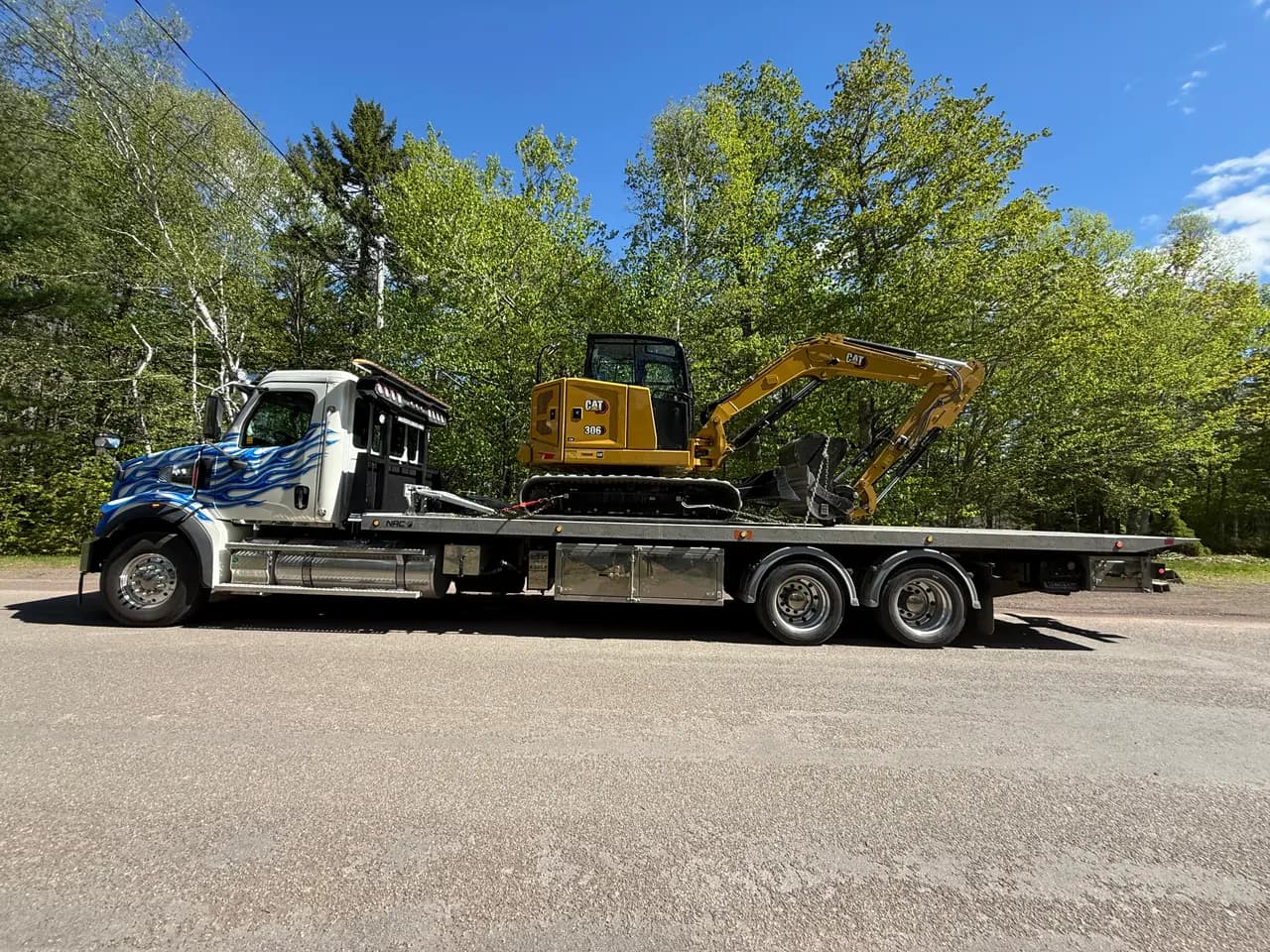 Dynamic Towing flat deck hauling a yellow CAT mini excavator on a New Brunswick road